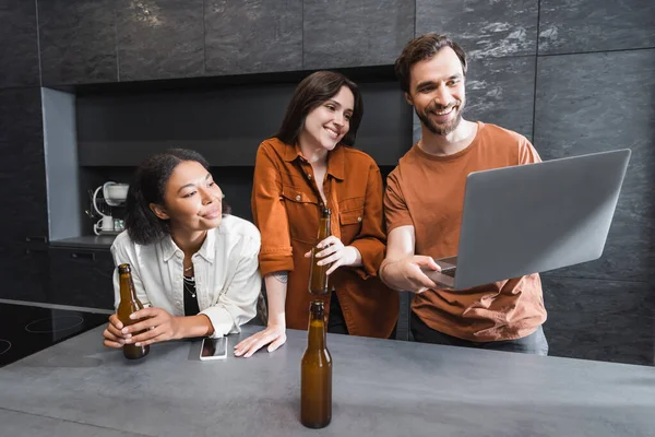 Счастливый человек держит ноутбук рядом с многонациональными друзьями с бутылками пива на кухне — стоковое фото