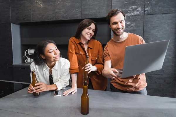 Hombre feliz sosteniendo portátil cerca de amigos multiétnicos con botellas de cerveza en la cocina - foto de stock