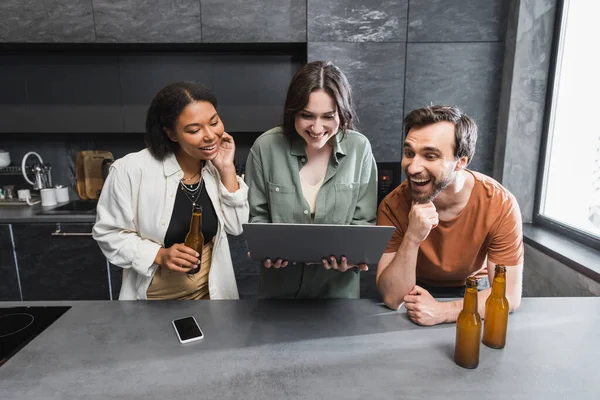 Donna felice che tiene il computer portatile vicino ad amici multietnici con bottiglie di birra in cucina — Foto stock