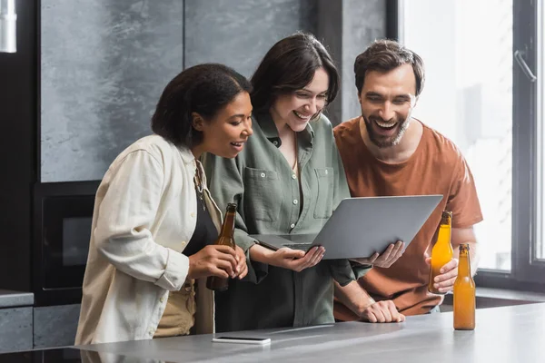 Mujer feliz sosteniendo portátil cerca de amigos interracial con botellas de cerveza en la cocina - foto de stock
