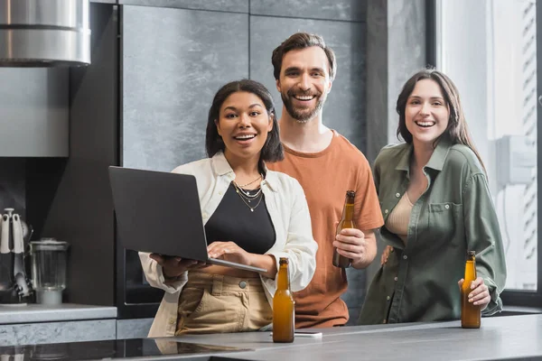 Счастливая двурасовая женщина держит ноутбук рядом с друзьями с бутылками пива, стоящими на кухне — стоковое фото