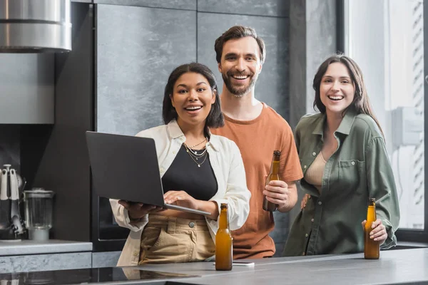 Felice donna bi-razziale che tiene il computer portatile vicino agli amici con bottiglie di birra in piedi in cucina — Foto stock