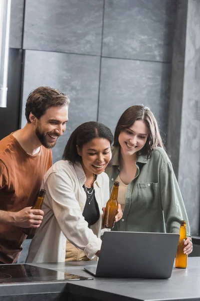 Amigos inter-raciais felizes segurando garrafas de cerveja e assistindo filme de comédia no laptop — Fotografia de Stock