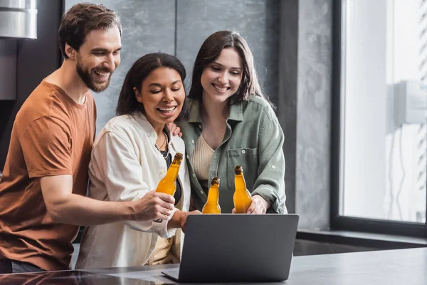 Allegri amici multietnici che tengono bottiglie di birra e guardano film comici sul computer portatile — Foto stock