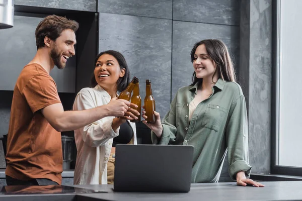 Alegres amigos multiétnicos sosteniendo botellas de cerveza y tintineo cerca del ordenador portátil en la cocina - foto de stock