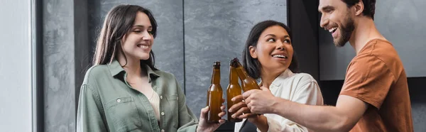 Fröhliche multiethnische Freunde, die Bierflaschen in der Hand halten und in der Küche klirren, Banner — Stockfoto