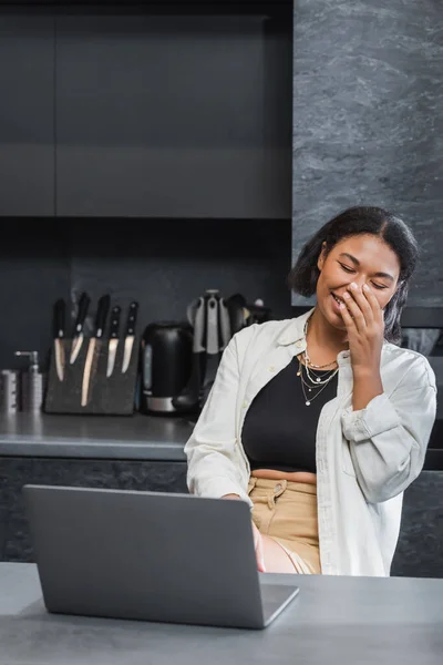 Heureuse femme bi-raciale couvrant la bouche et riant près d'un ordinateur portable dans la cuisine — Photo de stock