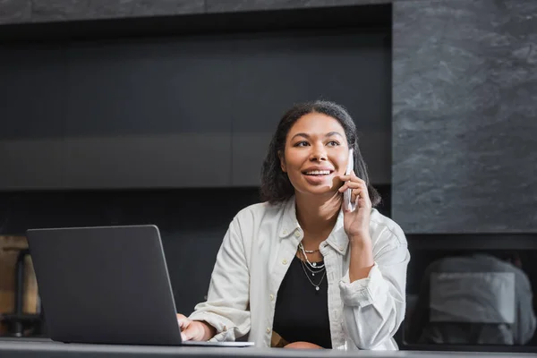 Happy bi-racial woman talking on smartphone near laptop on kitchen worktop — Stock Photo