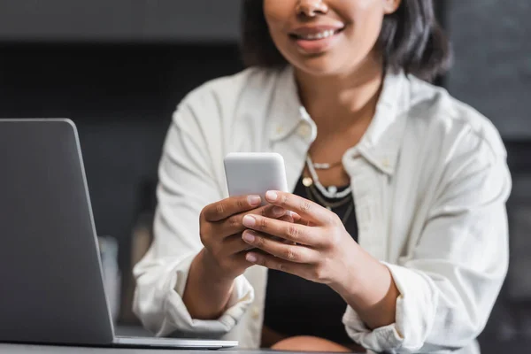 Vista recortada de mensajes de mujer bi-racial feliz en el teléfono inteligente cerca de la computadora portátil - foto de stock