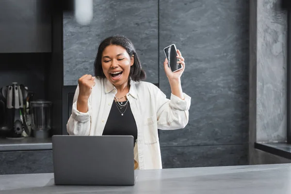 Excited bi-racial woman holding smartphone and looking at laptop in kitchen — Stock Photo