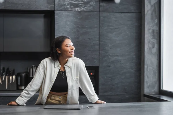 Excitée femme bi-raciale souriant tout en regardant loin près du smartphone et ordinateur portable sur le plan de travail de la cuisine — Photo de stock