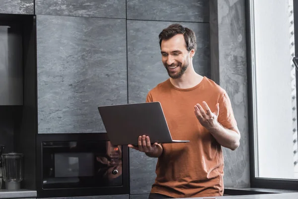 Homme barbu joyeux regardant ordinateur portable et gestuelle pendant l'appel vidéo — Photo de stock