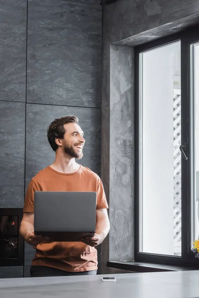 Happy bearded freelancer holding laptop while standing near smartphone on kitchen worktop — Stock Photo