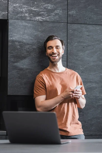 Happy bearded freelancer holding smartphone and looking at laptop — Stock Photo