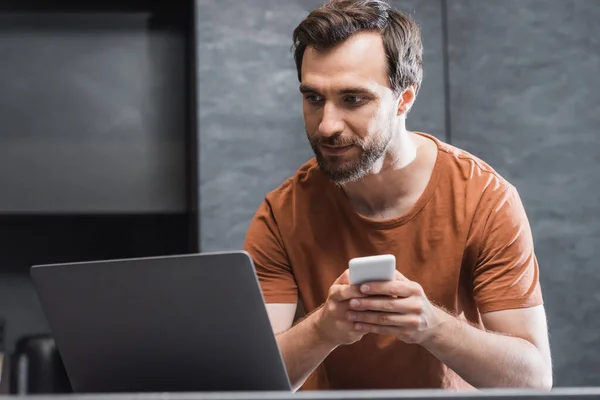 Bearded freelancer holding smartphone while looking at laptop — Stock Photo