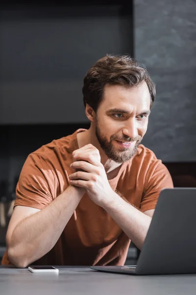 Alegre barbudo freelancer mirando portátil cerca de teléfono inteligente en encimera de cocina - foto de stock