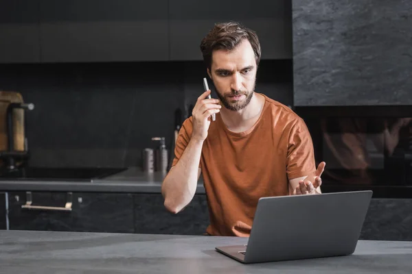 Serious bearded freelancer talking on smartphone and looking at laptop — Stock Photo