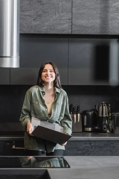 Cheerful young woman in green shirt holding laptop and looking at camera in kitchen — Stock Photo