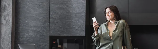 Cheerful young woman in green shirt holding smartphone near laptop in kitchen, banner — Stock Photo