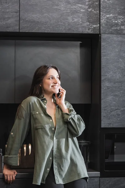 Joyeuse jeune femme en chemise verte parlant sur téléphone portable dans la cuisine — Photo de stock