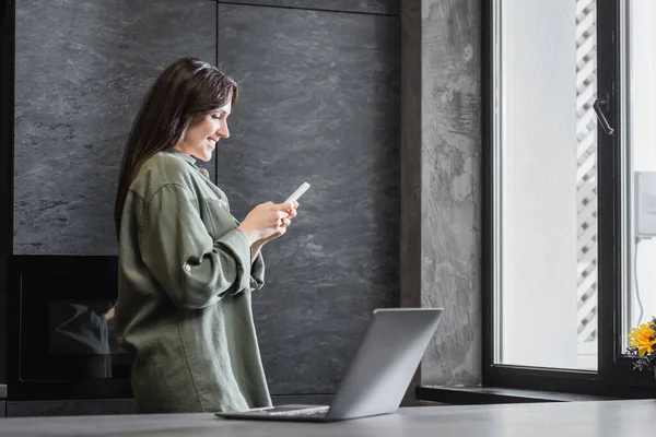 Fröhliche junge Freiberuflerin im grünen Hemd hält Smartphone neben Laptop auf Küchenarbeitsplatte — Stockfoto