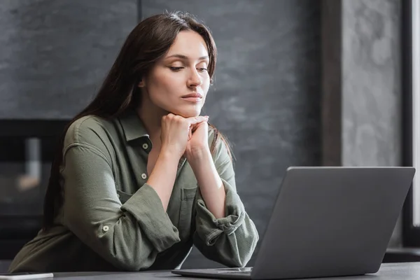 Mujer joven en camisa verde mirando a la computadora portátil mientras trabaja desde casa - foto de stock