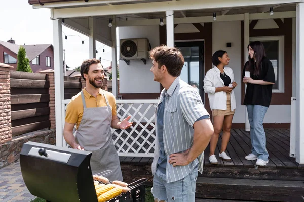 Cheerful men making barbecue and talking near blurred interracial friends on backyard — Stock Photo