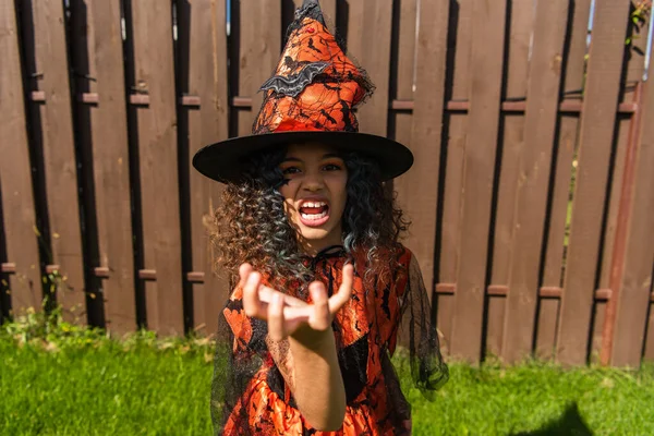 Chica en traje de bruja y sombrero puntiagudo mostrando gesto de miedo y gruñendo cerca de valla de madera - foto de stock