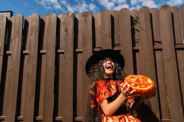 Ragazza eccitata in costume strega Halloween tenendo jack o lanterna e ridendo vicino alla recinzione di legno — Foto stock