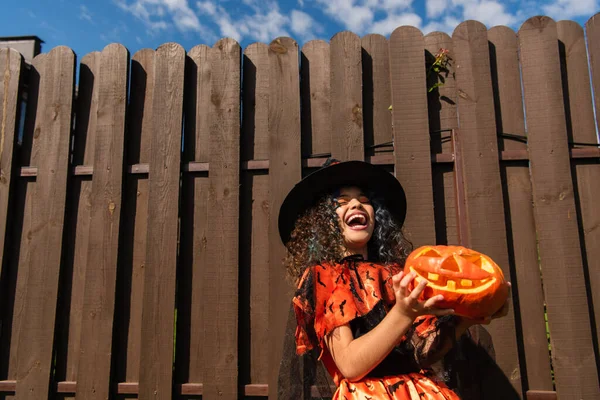Excited girl in witch halloween costume holding jack o lantern and laughing near wooden fence — Stock Photo