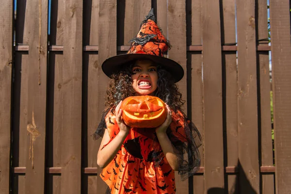 Girl in witch costume holding jack o lantern and showing scary grimace — Stock Photo