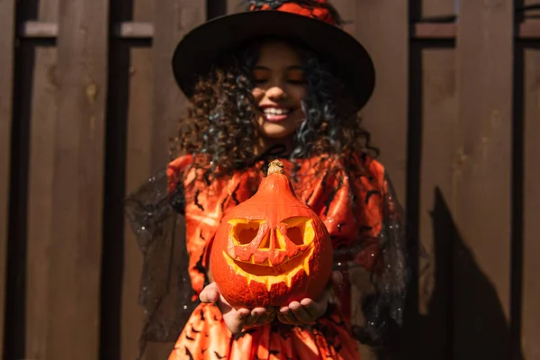 Enfoque selectivo de calabaza tallada en las manos de la chica sonriente en traje de Halloween bruja - foto de stock