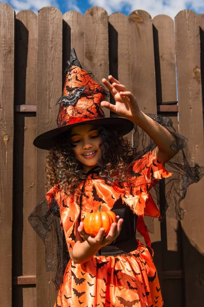 Smiling girl in halloween costume and witch hat gesturing near small pumpkin — Stock Photo