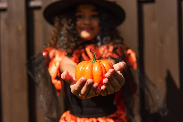 Selective focus of small pumpkin in hands of blurred girl in halloween costume — Stock Photo