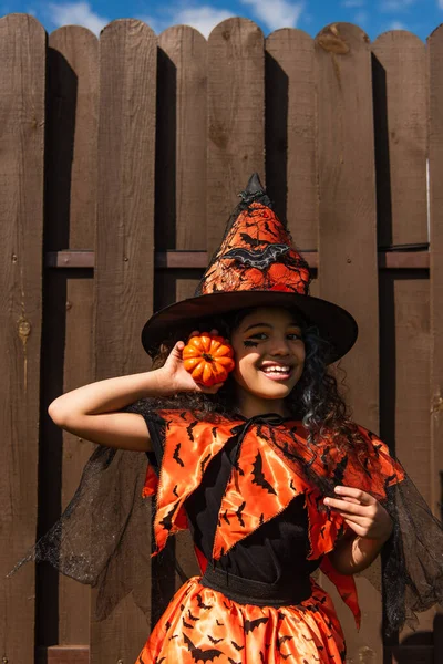 Cheerful curly girl in halloween costume and witch hat holding small pumpkin and looking at camera — Stock Photo