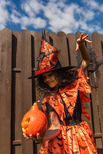 Smiling girl in witch costume and pointed hat gesturing near carved pumpkin — Stock Photo