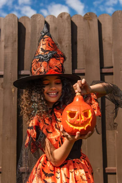Heureuse fille bouclée en costume d'Halloween et chapeau de sorcière tenant citrouille sculptée — Photo de stock