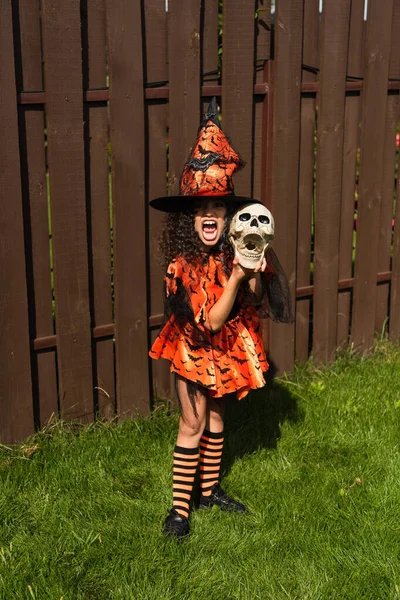 Pleine longueur de fille en costume d'Halloween et chapeau de sorcière tenant le crâne effrayant et grimaçant à l'extérieur — Photo de stock