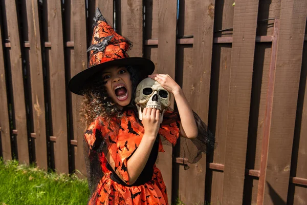 Criança em trajes de bruxa segurando o crânio assustador, sorrindo e rosnando ao ar livre — Fotografia de Stock