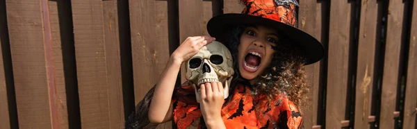 Fille en costume de sorcière tenant le crâne et grognant avec grimace en colère, bannière — Photo de stock