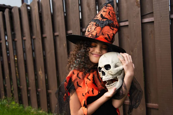 Glückliches Mädchen mit Hexenhut und Halloween-Kostüm, das gruseligen Totenkopf umarmt — Stockfoto