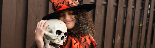 Pleased girl in witch hat embracing creepy skull outdoors, banner — Stock Photo