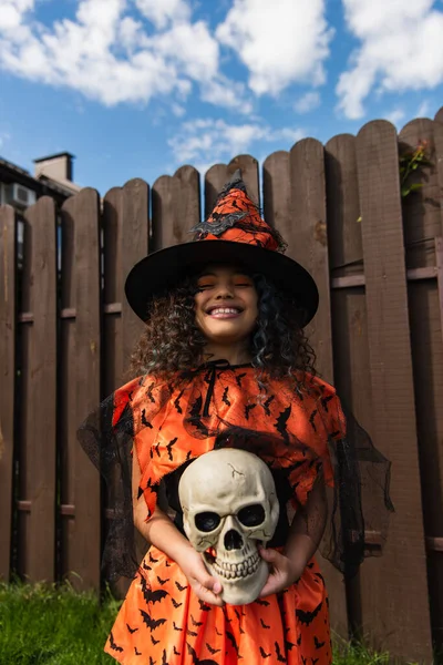 Menina sorridente em traje de bruxa e chapéu pontiagudo de pé com olhos fechados e crânio assustador — Fotografia de Stock