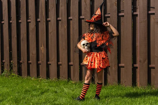Full length of girl in halloween costume and striped knee socks holding skull near wooden fence — Stock Photo