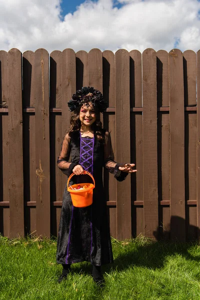 Chica sonriente en traje de bruja sosteniendo araña de juguete y cubo de halloween con dulces - foto de stock