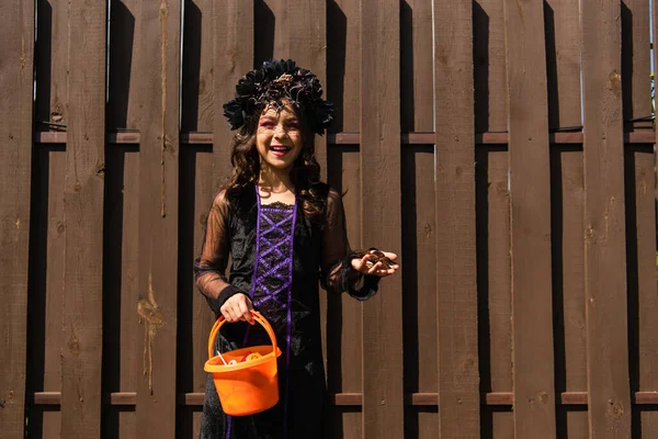 Menina alegre no traje de bruxa segurando aranha de brinquedo e truque ou tratar balde com doces — Fotografia de Stock