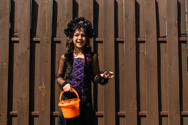 Alegre chica en bruja traje celebración juguete araña y truco o tratar cubo con dulces - foto de stock