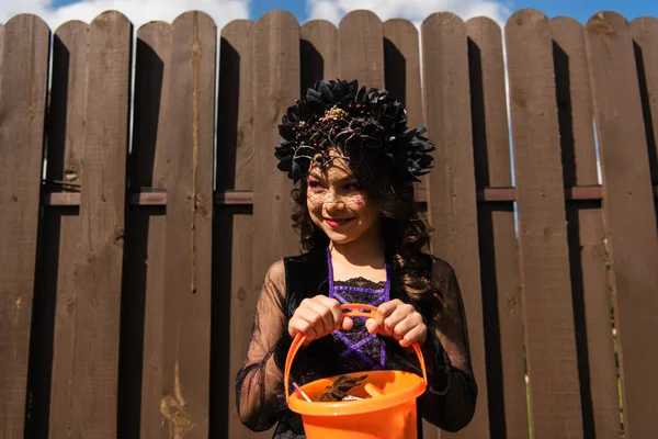Chica sonriente en corona negra sosteniendo cubo de halloween con dulces y mirando al aire libre - foto de stock