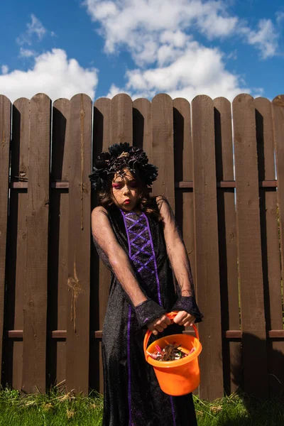Chica en traje de bruja y corona negra sosteniendo cubo pesado con caramelos - foto de stock