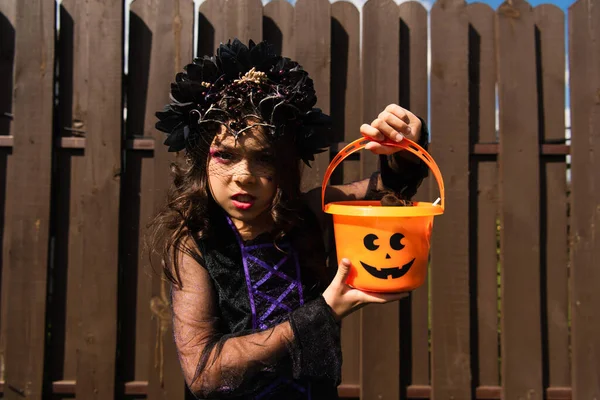 Chica en traje de bruja sosteniendo cubo de Halloween y mirando a la cámara con mueca enojada - foto de stock